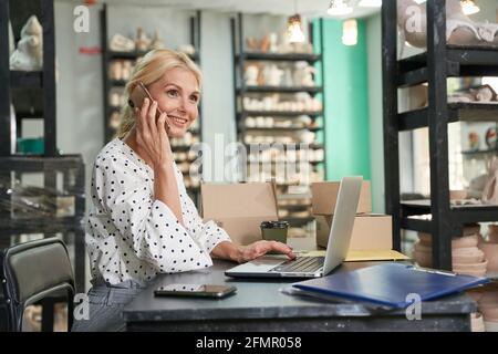 Selbstbewusste, reife Frau, Inhaberin eines Kleinunternehmens, die lächelt, während sie am Telefon spricht, ihren Laptop benutzt, Bestellungen entgegennimmt und in ihrem Handwerk am Schreibtisch sitzt Stockfoto