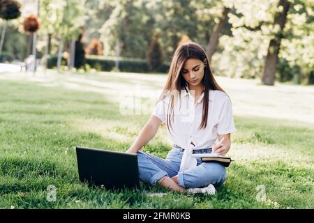 Schöne junge Studentin mit einem Laptop im Freien sitzt auf dem Gras, die Vorbereitung für Prüfungen. Technologie-, Bildungs- und Fernarbeitskonzept. Softselect Stockfoto