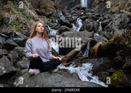 Mädchen sitzt auf einem großen Stein und meditiert in der Nähe eines Wasserfalls neben einem Bergfluss. Stockfoto