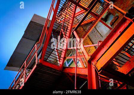 Von unten befindet sich eine rote Metalltreppe, die sich außerhalb des verwitterten Gebäudes mit befindet Ziegelmauer gegen wolkenlosen blauen Himmel Stockfoto