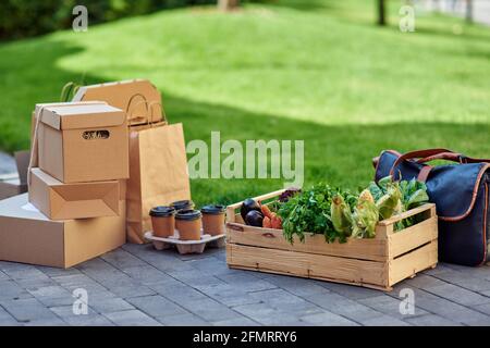 Holzkiste mit frischem Lebensmittelgeschäft, vier Pappbecher, verschiedene Pakete und Boxen mit Lebensmitteln, die draußen auf der Straße stehen Stockfoto