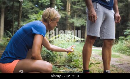 Ein Mann in Sportkleidung steht und eine Frau hockt sich in seiner Nähe und wendet Zecken- und Mückenschutz an, Spray, auf seine Beine. Stockfoto