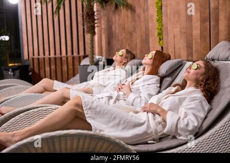 Zwei Frauen machen im Spa Schönheitsmasken. Gurken für Frische der Haut um die Augen. Ruhige Damen kümmern sich um Jugendliche Haut. Freundinnen liegen auf dem Bett in s Stockfoto
