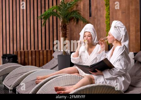 Zwei attraktive, entspannte Frauen in Bademänteln, die das Menü im Spa-Salon betrachten, Verfahren auswählen, sich ausruhen, kommunizieren Stockfoto