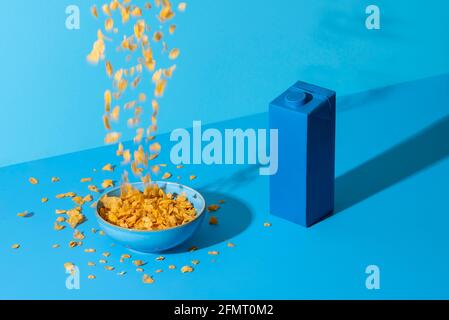 In eine Schüssel auf einen blauen Tisch gießen. Frühstückstisch mit Cornflakes, Müsli in einer Schüssel und einer Milchbox, in hellem Licht. Stockfoto