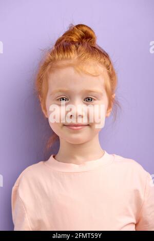 Nettes lächelndes kaukasisches Mädchen mit roten Haaren und großen blauen Augen isoliert über lila Hintergrund. Natürliche rothaarige Mädchen mit Sommersprossen Blick auf Kamer Stockfoto