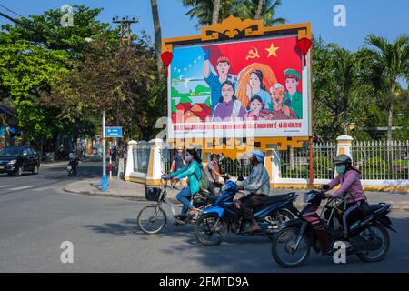 Vietnamesische Motorräder und Fahrräder warten an der Kreuzung vor dem kommunistischen Propagandaplakat Hoi an, Vietnam Stockfoto