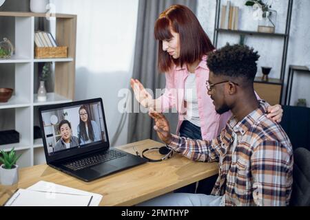 Glücklicher afrikanischer Mann und kaukasische Frau lächeln und winken während des Videochats mit ihren asiatischen Freunden. Junges Paar, das zu Hause bleibt und einen modernen Laptop für die Kommunikation nutzt. Stockfoto