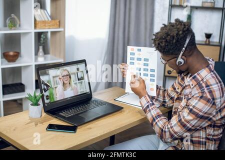 Seitenansicht eines afro-amerikanischen Studenten, der während der Pandemie-Zeit die Prüfungen durch Videoanruf auf einem modernen Laptop abgibt. Kaukasische Lehrerin mit blonden Haaren auf dem Computerbildschirm. Stockfoto