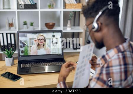 Seitenansicht eines afro-amerikanischen Studenten, der während der Pandemie-Zeit die Prüfungen durch Videoanruf auf einem modernen Laptop abgibt. Kaukasische Lehrerin mit blonden Haaren auf dem Computerbildschirm. Stockfoto