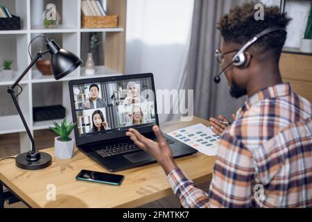 Multiethnische Gruppe von Personen, die Geschäftstreffen durch Videoanruf auf einem Laptop abhalten. Blick von der Schulter auf einen schwarzen Mann, der im Heimbüro sitzt und aus der Ferne arbeitet. Stockfoto