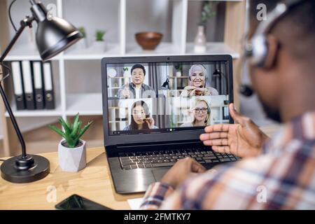 Multiethnische Gruppe von Personen, die Geschäftstreffen durch Videoanruf auf einem Laptop abhalten. Blick von der Schulter auf einen schwarzen Mann, der im Heimbüro sitzt und aus der Ferne arbeitet. Stockfoto