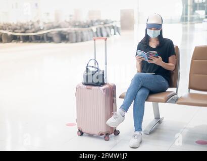 Asiatische Reisende, die auf dem internationalen Flughafen ein chirurgisches Zeichen mit Handgepäck trägt und während der Covid auf ihren Flug wartet, ein Buch liest Stockfoto