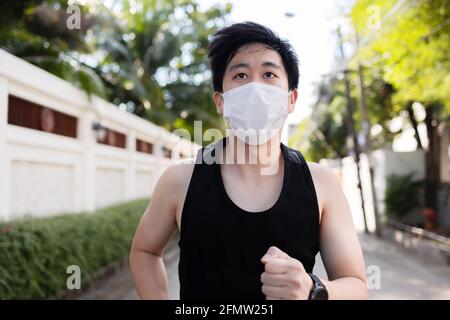 Junger asiatischer Mann, der mit einer Gesichtsmaske in der Stadt läuft. Laufmännchen trainieren und joggen auf der Straße Stockfoto