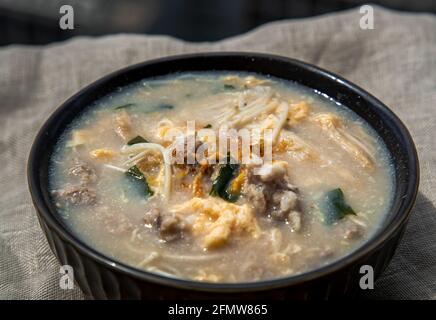 Nahaufnahme von Schweinefleisch-Congee oder chinesischem Reisbrei mit gehacktem Schweinefleisch, Ei, Enoki-Pilz und Algen in einer Schüssel auf weißem Tischset. Asiatisches Frühstück, Selecti Stockfoto