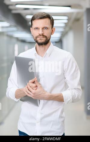 Porträt eines hübschen jungen Mannes, der einen Laptop in der Hand hält und fröhlich die Kamera anlächelt, den Platz zum Kopieren. Ein Mann im Hintergrund des Büros Stockfoto