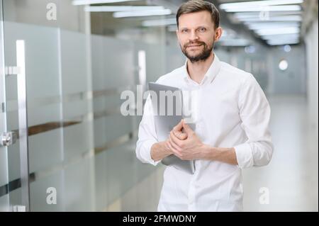 Porträt eines hübschen jungen Mannes, der einen Laptop in der Hand hält und fröhlich die Kamera anlächelt, den Platz zum Kopieren. Ein Mann im Hintergrund des Büros Stockfoto