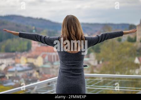 Frau, die auf einem Balkon mit ausgestreckten Armen in einem steht Rückansicht, als sie über eine Kirche und schaut Stadt unten mit Bergkulisse Stockfoto