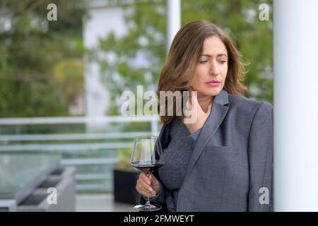 Frau, die sich auf dem Balkon bei einem Glas Wein entspannt Mit einer Jacke, die über sie drapiert ist, gegen das Dailing lehnend Schultern, die pensiv zur Seite schauen Stockfoto