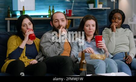Gemischte Freunde, die sich auf dem Sofa entspannen, während sie sich einen komischen Film im Fernsehen ansehen, und lachend während der Heimparty spät in der Nacht im Wohnzimmer. Gruppe von multirassischen Menschen genießen Zeit zusammen Stockfoto