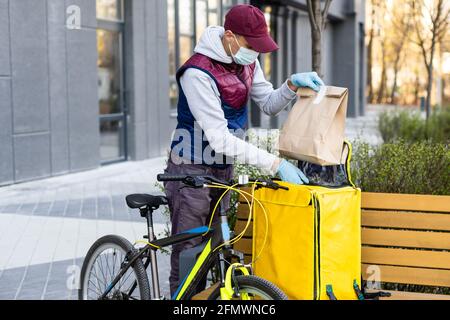 Junger Kurier in medizinischer Maske liefert Lebensmittel mit gelbem Thermalrucksack, fährt mit dem Fahrrad in der Stadt. Konzept für den Lieferservice von Lebensmitteln Stockfoto