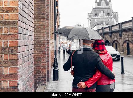 Ein Paar, das im Regen unter einer wunderschönen Tower Bridge von London spazieren geht. Rentnerpaar, das um die Welt reist. Rentner, Liebe und Urlaubskonzept. Stockfoto