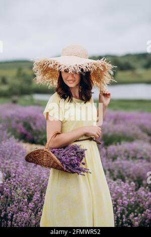 Porträt einer Frau in einem gelben Kleid und einem Strohhut mit einem Blumenkorb in einem Lavendelfeld. Weicher, selektiver Fokus, Bildrauschen Stockfoto