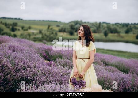 Junge schöne Frau in einem gelben Kleid mit einem Korb mit Blumen in einem Lavendelfeld. Weicher, selektiver Fokus, Bildrauschen Stockfoto