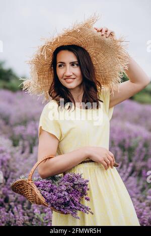 Porträt einer Frau in einem gelben Kleid und einem Strohhut mit einem Blumenkorb in einem Lavendelfeld. Weicher, selektiver Fokus, Bildrauschen Stockfoto