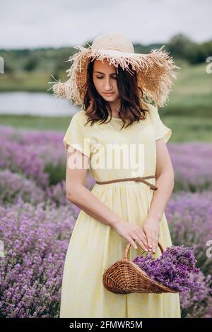 Porträt einer Frau in einem gelben Kleid und einem Strohhut mit einem Blumenkorb in einem Lavendelfeld. Weicher, selektiver Fokus, Bildrauschen Stockfoto