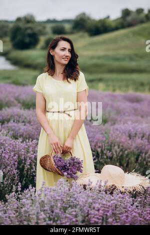 Junge schöne Frau in einem gelben Kleid mit einem Korb mit Blumen in einem Lavendelfeld. Weicher, selektiver Fokus, Bildrauschen Stockfoto