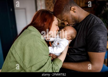 Glückliche multirassische gemischte Familie sanft halten neugeborenes Baby. Rothaarige kaukasische Mutter, die ihr Kind ansieht, afroamerikanischer Vater, der sich liebevoll niedlich küsst Stockfoto