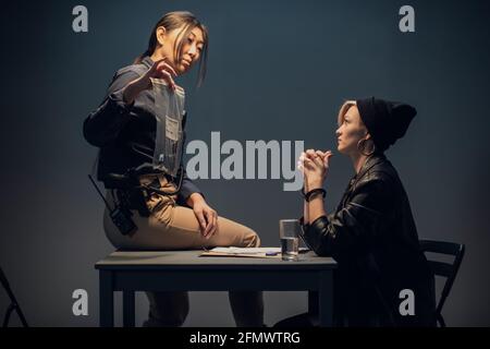 Eine junge asiatische Forscherin sitzt auf einem Tisch und zeigt einem blonden Verdächtigen eine Packung Waffen. Stockfoto