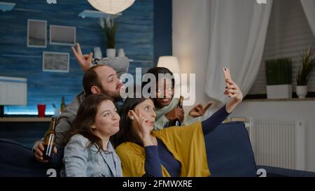 Multirassische Freunde, die sich auf dem Sofa ausruhen, verbringen gerne Zeit miteinander, während sie Selfie über soziale Medien teilen. Eine Gruppe multiethnischer Menschen, die spät in der Nacht im Wohnzimmer lachen Stockfoto