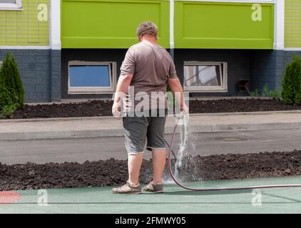 Mann, der in der Hitze vor seinem Haus den Boden herunterschwärmte. Mann wässert Boden mit Schlauchleitung. Gartenarbeit, Verbesserung der psychischen Gesundheit Stockfoto