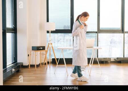 Porträt einer denkenden Ärztin im weißen Mantel, die an sonnigen Tagen auf dem Fensterhintergrund im hellen medizinischen Klinikbüro steht. Stockfoto