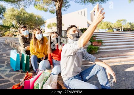 Gruppe von jungen Menschen in schützenden medizinischen Maske unter mehreren Farbige Einkaufstaschen warten auf den Einkaufsmarkt und genießen das Internet Mobilfunkverbindung t Stockfoto