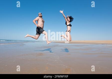 Junges Paar im Urlaub lacht und genießt beim Springen und Laufen an einem einsamen Strand. Freizeit- und Urlaubskonzept Stockfoto