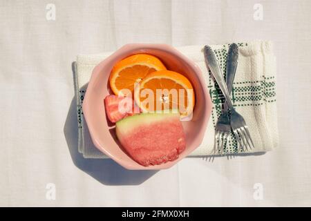 Detailbild in horizontaler Darstellung von verschiedenen Früchten (Orangen, Wassermelone und Erdbeere) in einer rosa Schale zusammen mit zwei Gabeln auf einem Handtuch in einem weißen Stoff Stockfoto
