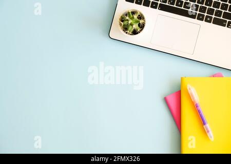 Büroarbeitsplatz mit Laptop, mehreren bunten Notizbüchern, grüner Kaktuspflanze, weißer Tasse Kaffee, Verbrauchsmaterial und Papeterie auf blauem Hintergrund. Schließen Stockfoto
