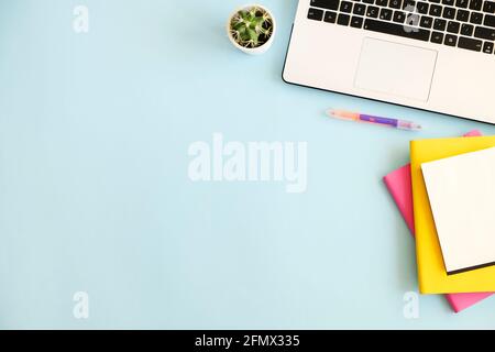 Büroarbeitsplatz mit Laptop, mehreren bunten Notizbüchern, grüner Kaktuspflanze, weißer Tasse Kaffee, Verbrauchsmaterial und Papeterie auf blauem Hintergrund. Schließen Stockfoto