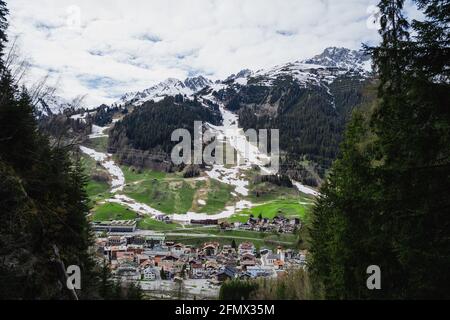 Weite Aussicht über einen klaren Bach, ein winziges Dorf und schneebedeckte Berge. Die Aussicht ist von einem Hügel aus aufgenommen. Das Bild wird von einem Nadelbaumholz umrahmt. Stockfoto