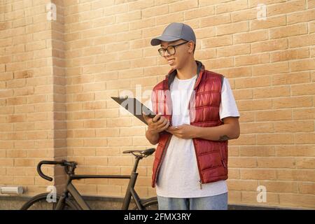 Junger asiatischer männlicher Kurier, der im Stehen auf Clipboard schaut Stockfoto