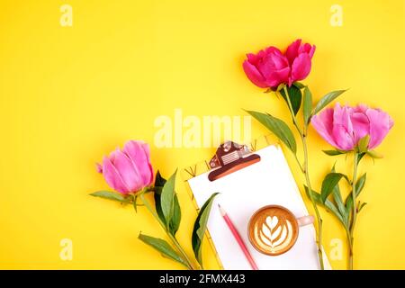 Frauenarbeitsplatz, leeres Papierblatt auf einem Klemmbrett, Tasse Kaffee, pinker Stift & lilafarbene Pfingstrosen-Blumen auf gelbem Tischhintergrund. Feminine flache Laie c Stockfoto