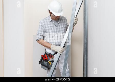 Man construction worker or plasterer holding drywall metal profiles near plasterboard white wall in building site. Wearing white hardhat, work gloves, Stockfoto