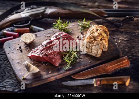 Nährstoffreiches, rohes Hirschwild, das auf einem rustikalen Holztisch mit geröstetem Knoblauch, Rosmarin und hungriges Zubehör wie Shot gu für den Kochvorgang zubereitet wird Stockfoto