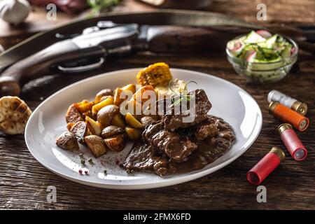 Wildgulasch-Eintopf, serviert mit gerösteten Kartoffeln und gewürzt mit vierfarbigem Pfeffermais. Das ganze Essen ist von Jagdflinte und Kanonenbül umgeben Stockfoto