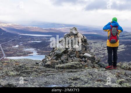 Nach einem Regen im Spätherbst steht ein Mann auf dem Gipfel von Mehøe neben einem Steinhaufen und blickt in das Tal mit seinen Seen. Dovre-Nationalpark Stockfoto