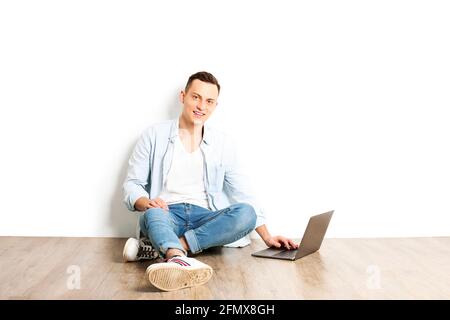Porträt eines glücklichen jungen Mannes mit einem Lächeln in einem weißen Hemd ohne Knöpfe, der auf einem Hartholzboden mit einem Laptop sitzt. Selbstbewusster lässiger Hipster Stockfoto