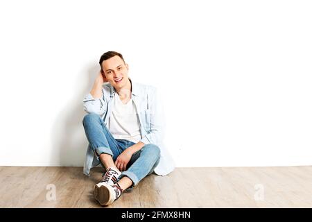 Porträt eines glücklichen jungen Millennials mit strahlenden Lächeln in einem weißen Hemd ohne Knöpfe sitzt auf einem Holzboden, isoliert. Selbstbewusster lässiger Hippie-Look Stockfoto